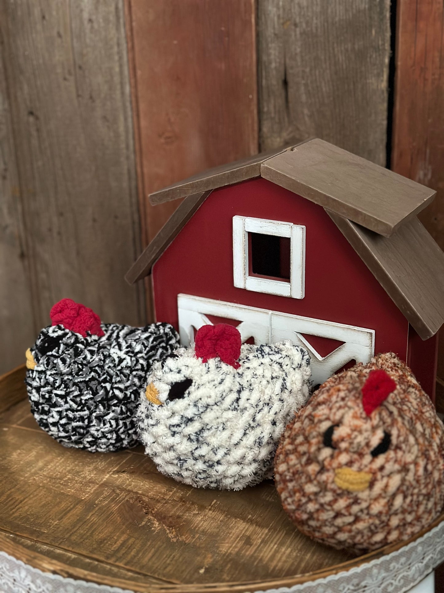 crocheted chicken stufies in front of a small barn model on a wooden surface.