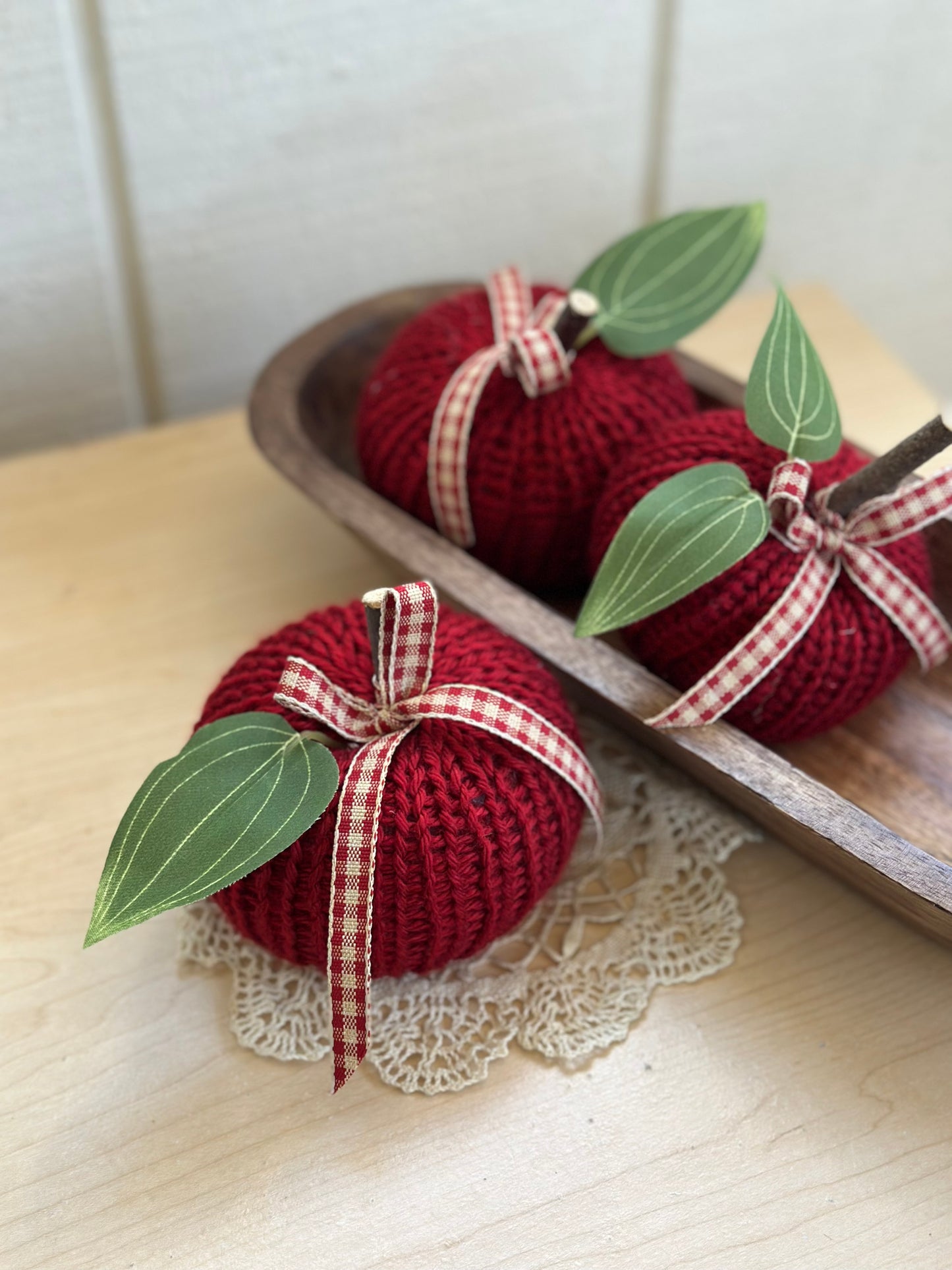 Knitted red apples with green leaves and red checkered ribbons on a wooden tray.