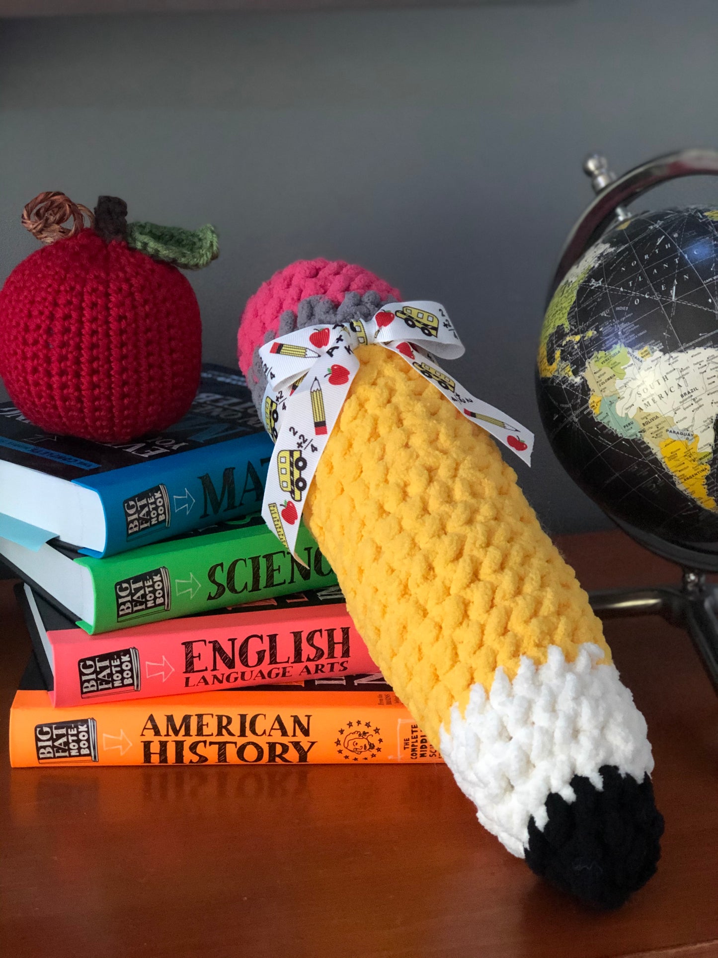 Crocheted pencil with playing cards, apple, and books on a desk with a globe in the background