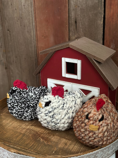crocheted chicken stufies in front of a small barn model on a wooden surface.