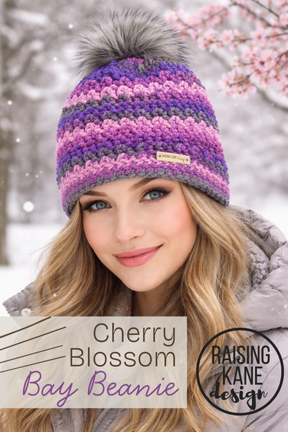 Woman wearing a colorful striped beanie with a fluffy pom-pom in a snowy setting, with 'Raising Kane Design' branding. photo is AI generated.