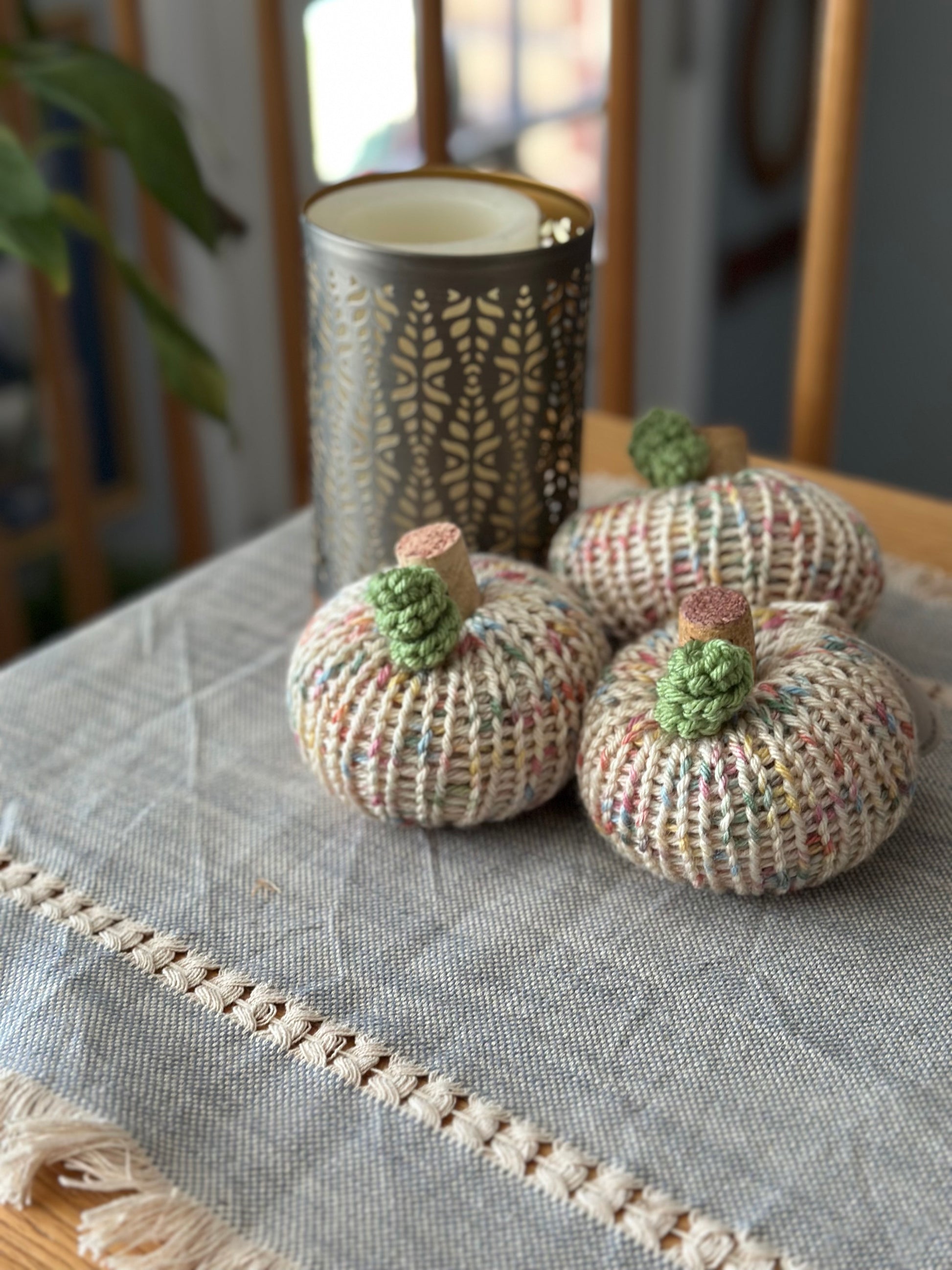 Three knitted pumpkins on a textured tablecloth with a decorative vase in the background.